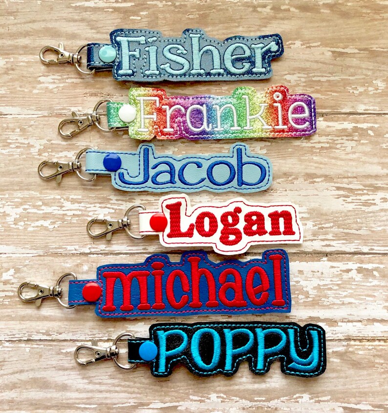 Pode incluir: Seis chaveiros personalizados com diferentes nomes bordados. Os nomes s&atilde;o Fisher, Frankie, Jacob, Logan, Michael e Poppy. Cada chaveiro tem uma cor e um design diferente.