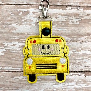 May include: A yellow school bus keychain with a smiling face, black wheels, and a silver clip.
