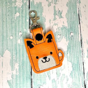 May include: Orange fox-shaped leather keychain with black ears and a white embroidered face. The keychain has a silver metal ring and a silver metal clip.