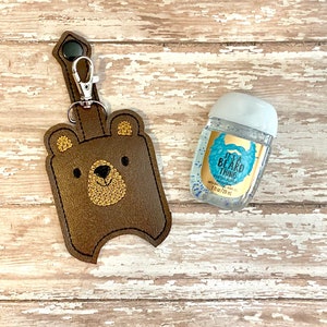 May include: Brown leather bear-shaped hand sanitizer holder with a gold embroidered face and a silver keychain. The holder is attached to a clear plastic bottle of hand sanitizer with a blue and gold label that reads "It's a Beard Thing" and "Antibacterial Hand Gel".