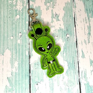 May include: Green glitter alien keychain with a winking face and a black belt. The keychain has a silver metal clip.