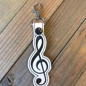 May include: White leather keychain with a black embroidered treble clef and a black button. The keychain has a silver metal key ring.