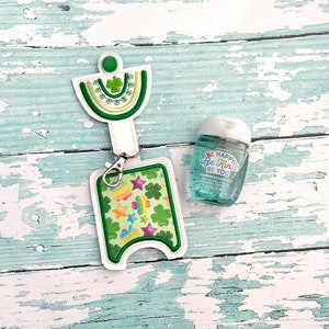 May include: A green and white hand sanitizer holder with a shamrock design and a rainbow and star pattern. The holder has a key ring and is attached to a small bottle of hand sanitizer that says "Be Happy Be Kind Be You".