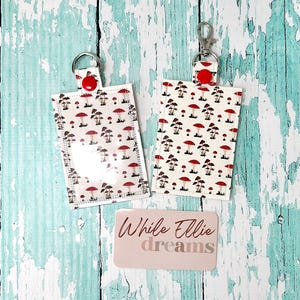 May include: Two white fabric keychains with a red mushroom pattern and clear plastic windows. The keychains have silver key rings and red button closures. The text "While Ellie dreams" is printed on a pink card below the keychains.