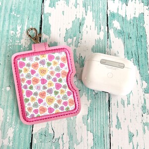 May include: A pink AirPod case holder with a silver clasp, featuring a Valentine's Day candy heart pattern. The case is embroidered with a pink border and holds a white AirPod case.