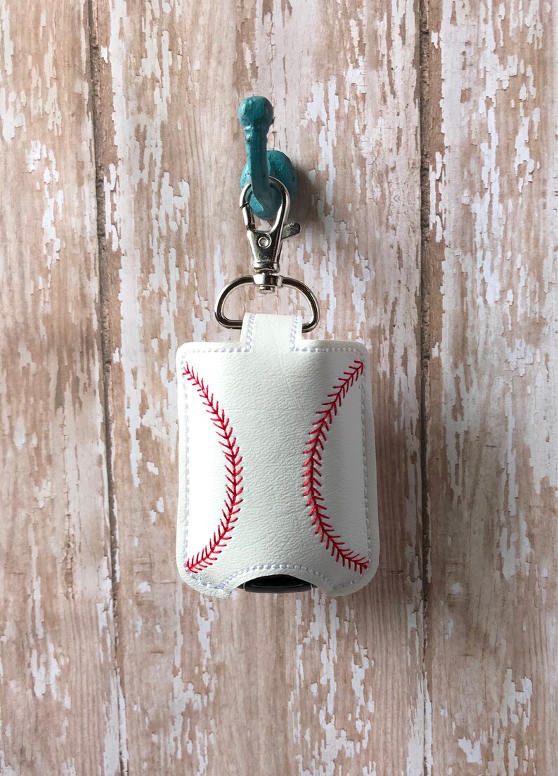 May include: White leather hand sanitiser holder with a red baseball stitch design. The holder is attached to a silver key chain and hangs from a turquoise hook.
