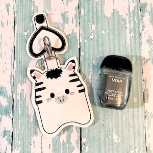 Pode incluir: Um porta-gel antibacteriano com design de zebra em branco e preto com um chaveiro em forma de coração. O porta-gel está ligado a uma pequena garrafa transparente de gel antibacteriano etiquetada "Men's Collection Noir Pocketbac Anti-Bacterial Hand Gel 1 fl oz/29 ml."
