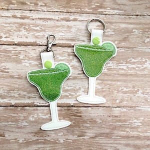 May include: Two green margarita glass keychains with lime wedges. The keychains are made of felt and have a sparkly green finish.