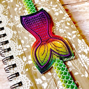 Puede incluir: Un marcap&aacute;ginas de cola de sirena arco&iacute;ris con un acabado iridiscente verde, morado y naranja. El marcap&aacute;ginas est&aacute; unido a un cuaderno con un dise&ntilde;o de encaje blanco.