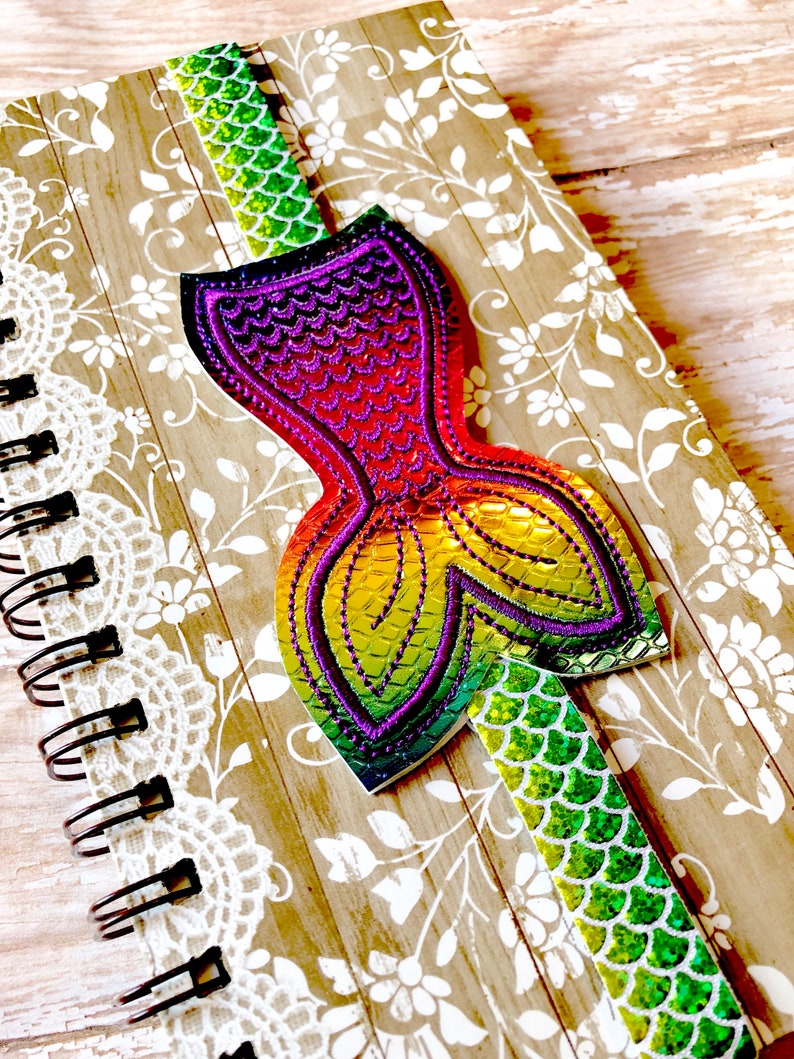 Puede incluir: Un marcap&aacute;ginas de cola de sirena arco&iacute;ris con un acabado iridiscente verde, morado y naranja. El marcap&aacute;ginas est&aacute; unido a un cuaderno con un dise&ntilde;o de encaje blanco.