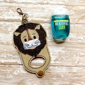 May include: A brown lion-shaped hand sanitizer holder with a glitter finish. The holder has a small loop for attaching a keychain. A blue hand sanitizer bottle with the text "PocketBac Bath & Body Works Beautiful Day Anti-Bacterial Hand Sanitizer 1 fl oz/29 ml" is visible in the background.