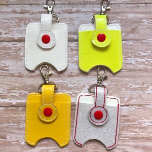 May include: Four hand sanitiser holders with keychains. The holders are made of leather and come in white, yellow, neon yellow, and grey. Each holder has a red button and a white stitching detail.