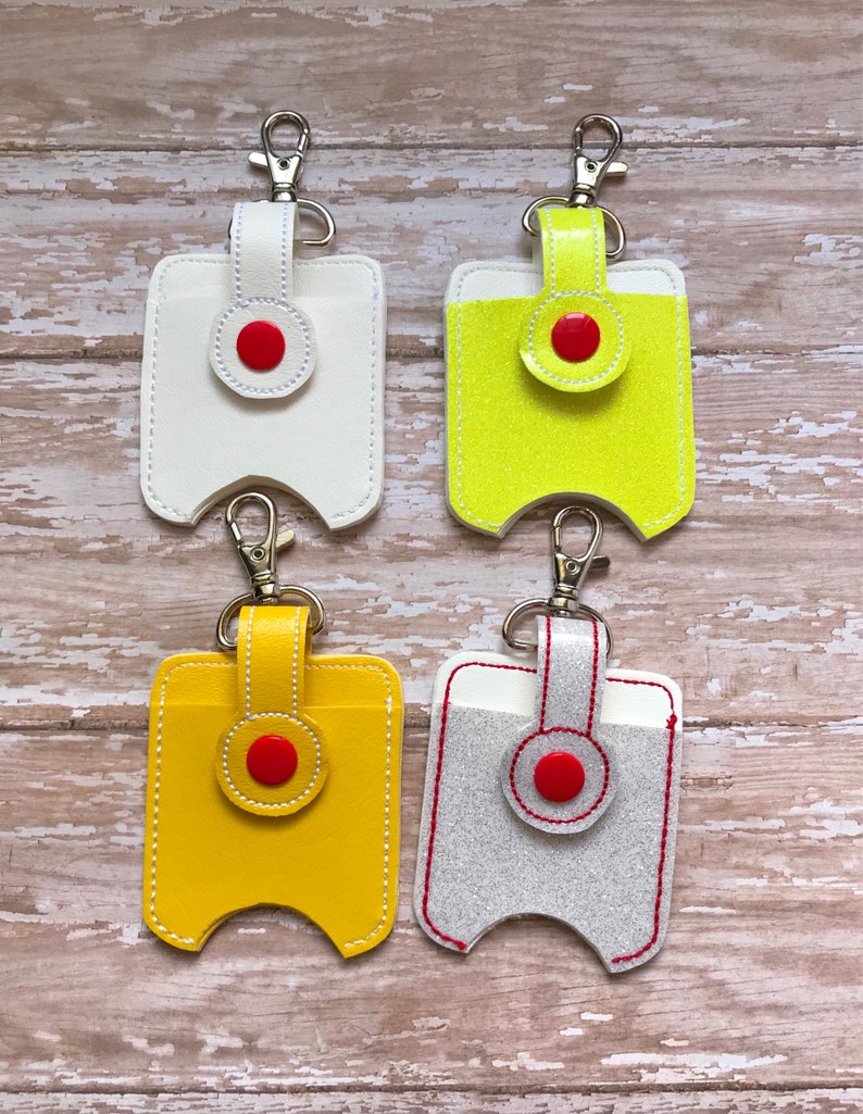 May include: Four hand sanitiser holders with keychains. The holders are made of leather and come in white, yellow, neon yellow, and grey. Each holder has a red button and a white stitching detail.