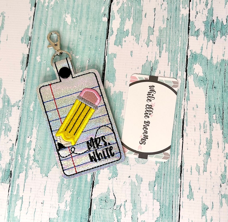 May include: A silver glitter keychain with a yellow pencil and the text "Mrs. White" on a white and gray notebook paper background.