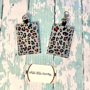 May include: Two keychains with a leopard print fabric and rainbow accents. The keychains are attached to a silver key ring. The text "while ellie dreams" is visible on a card in the background.
