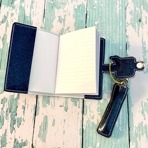 May include: A small, blue denim notebook with a pen holder attached. The notebook is open to reveal lined pages. The pen holder is made of denim and has a metal chain.