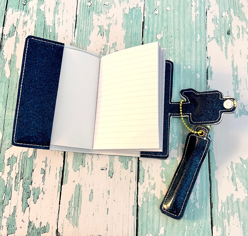 May include: A small, blue denim notebook with a pen holder attached. The notebook is open to reveal lined pages. The pen holder is made of denim and has a metal chain.