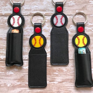 May include: Four black leather keychains with a baseball or softball design. Each keychain has a small pocket for a lip balm or other small item. The keychains are decorated with red, white, and yellow stitching.