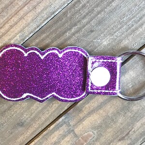 May include: Purple glitter keychain with a white outline and a silver key ring. The keychain has a white button on the back.
