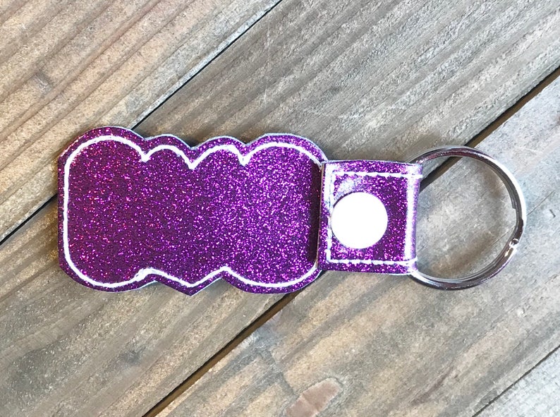 May include: Purple glitter keychain with a white outline and a silver key ring. The keychain has a white button on the back.