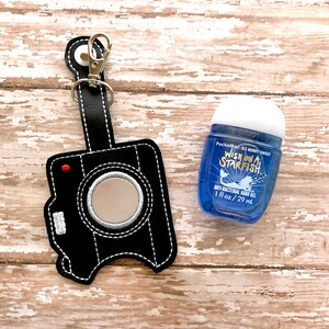 May include: Black camera-shaped keychain with a small mirror and a silver clip. The keychain is holding a blue bottle of hand sanitizer with the text "Pocketbac So Berry Sweet Wish On A Starfish Anti-Bacterial Hand Gel 1 fl oz/29 ml."