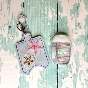 May include: A light blue hand sanitiser holder with a starfish and a sand dollar design. The holder has a silver keychain attached. A clear plastic bottle of hand sanitiser with the text "Fiji White Sands Anti-Bacterial Hand Gel 1 fl oz / 29 mL" is next to the holder.