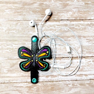 May include: A black butterfly-shaped headphone holder with a turquoise button. The holder is wrapped around white headphone cords.