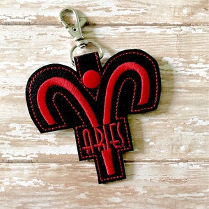 May include: Black and red embroidered keychain with the zodiac symbol for Aries and the word "Aries" in red thread.