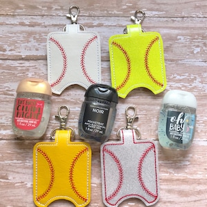 May include: Five baseball-themed hand sanitiser holders with keyrings. The holders are made of white, yellow, and grey fabric with red stitching. The holders are filled with hand sanitiser bottles. The bottles are labelled "But First, Champange", "Noir", and "Oh Baby".