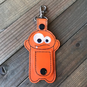 May include: Orange monster-shaped key chain with a pocket for a small item. The monster has white eyes and a black mouth with fangs.