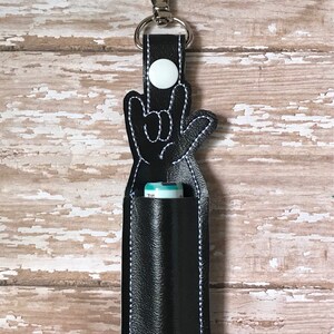 May include: Black leather lip balm holder with a white button and a hand-shaped design making the 'I love you' sign in American Sign Language.