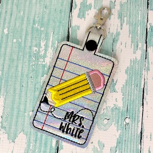 May include: A white key chain with a silver clip and a black button. The key chain has a design of a yellow pencil on a sheet of notebook paper with the text "Mrs. White" written on it.