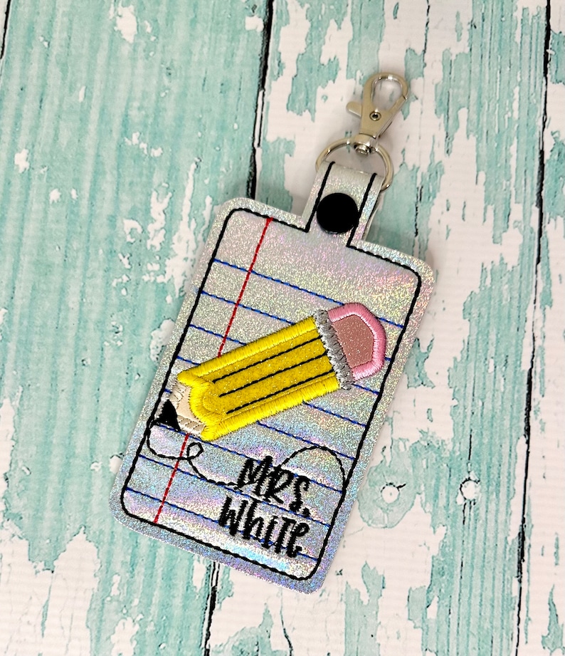 May include: A white key chain with a silver clip and a black button. The key chain has a design of a yellow pencil on a sheet of notebook paper with the text "Mrs. White" written on it.