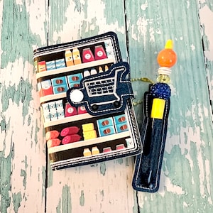 May include: A blue and black fabric notebook with a grocery store aisle design and a shopping cart on the front cover. The notebook is attached to a blue and yellow pen with a blue and orange bead charm.