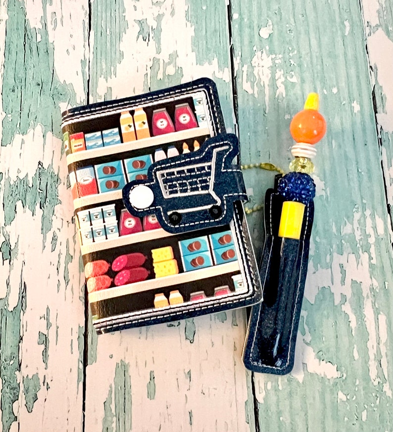 May include: A blue and black fabric notebook with a grocery store aisle design and a shopping cart on the front cover. The notebook is attached to a blue and yellow pen with a blue and orange bead charm.