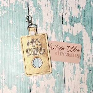 May include: Gold key chain with a clear plastic window and the text "MRS. BAIRD" embroidered in white thread. The key chain is on a wooden surface with a pink card that says "While Ellie dreams".