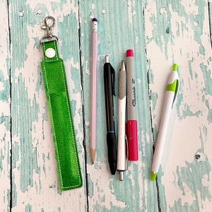 May include: A green leather keychain with a silver key ring and a white button. The keychain is lying on a light blue wooden surface with five writing utensils: a pink pencil, a black pen, a silver pen, a red marker, and a white marker.