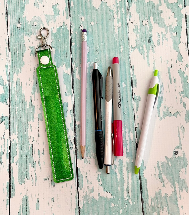 May include: A green leather keychain with a silver key ring and a white button. The keychain is lying on a light blue wooden surface with five writing utensils: a pink pencil, a black pen, a silver pen, a red marker, and a white marker.