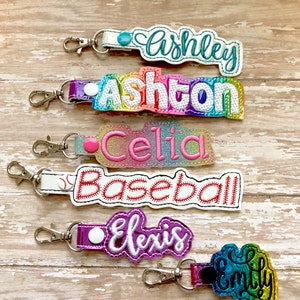 May include: Five colorful personalized keychains with different names and designs. The keychains feature names like Ashley, Ashton, Celia, Alexis, and Emily. Some keychains have a rainbow design, while others have a baseball or glitter design.