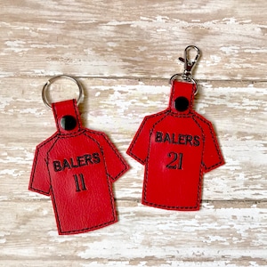 May include: Two red leather keychains shaped like baseball jerseys with black stitching. The jerseys have the word "BALERS" and a number, one with the number "11" and the other with the number "21".