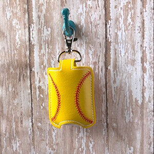 May include: Yellow softball-shaped hand sanitiser holder with red stitching. The holder is attached to a silver keychain.