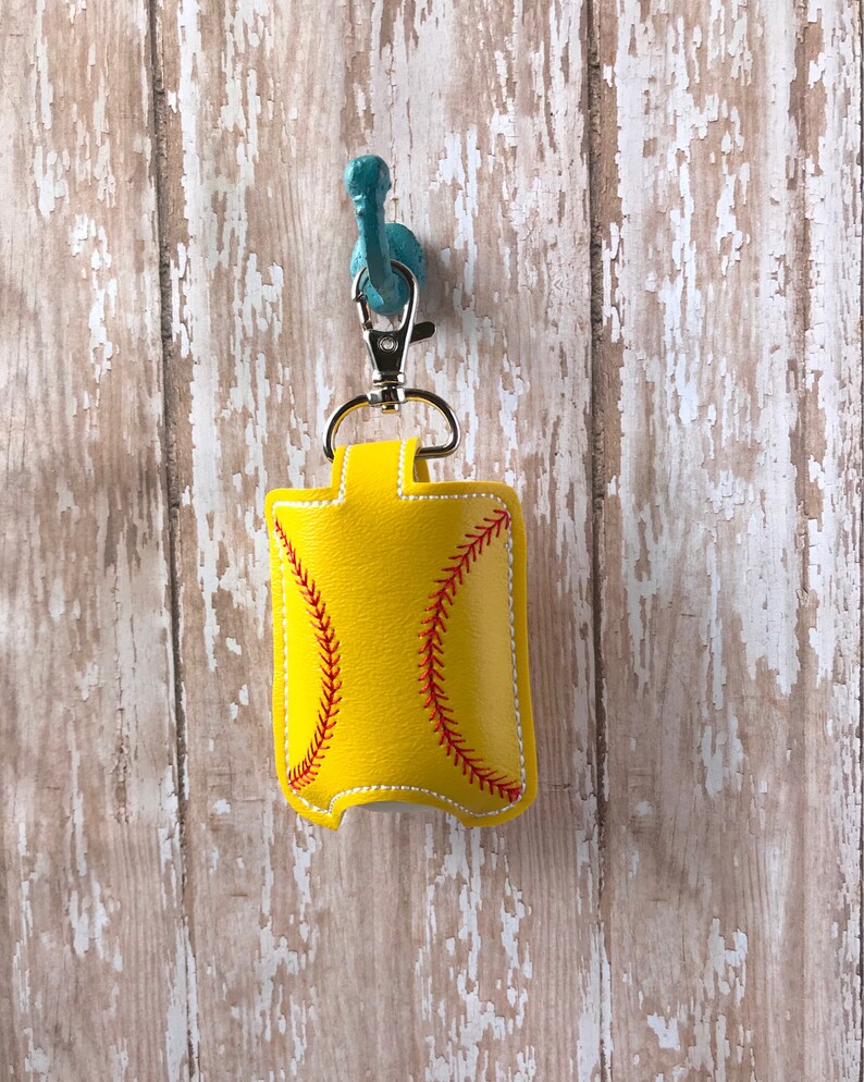 May include: Yellow softball-shaped hand sanitiser holder with red stitching. The holder is attached to a silver keychain.