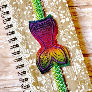 Puede incluir: Un marcap&aacute;ginas de cola de sirena colorido con escamas iridiscentes. El marcap&aacute;ginas est&aacute; unido a un cuaderno de espiral con un dise&ntilde;o floral.