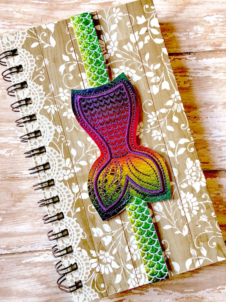 Puede incluir: Un marcap&aacute;ginas de cola de sirena colorido con escamas iridiscentes. El marcap&aacute;ginas est&aacute; unido a un cuaderno de espiral con un dise&ntilde;o floral.
