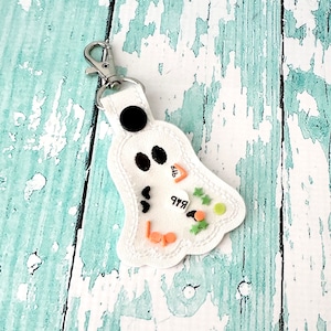 May include: White ghost-shaped keychain with black eyes and mouth. The keychain has a silver clasp and a black button. The ghost has orange and green embellishments and the word "Boo" in orange.