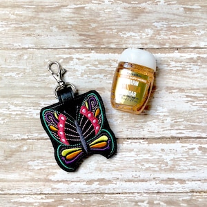 May include: A black butterfly-shaped keychain with colorful embroidery. The keychain is attached to a small bottle of PocketBac Warm Vanilla Sugar antibacterial hand gel.