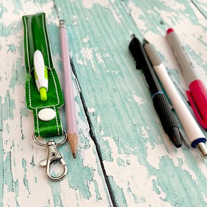 May include: A green fabric key chain with a silver clasp and a white circle. A green and white pen is inside the key chain. A pink pencil, a black pen, and a white pen with a pink cap are on a light blue wooden surface.