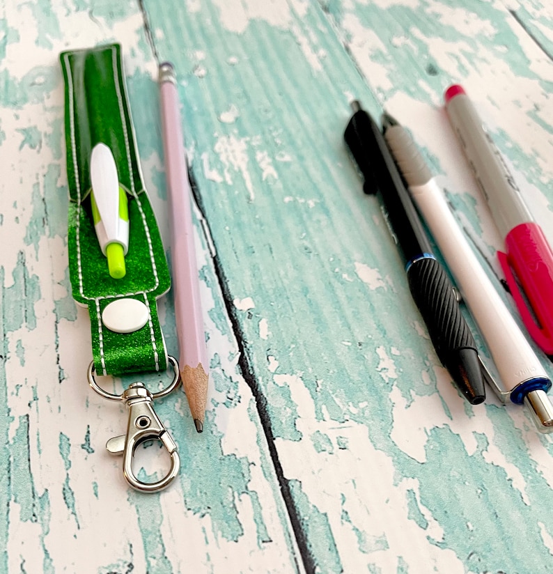 May include: A green fabric key chain with a silver clasp and a white circle. A green and white pen is inside the key chain. A pink pencil, a black pen, and a white pen with a pink cap are on a light blue wooden surface.
