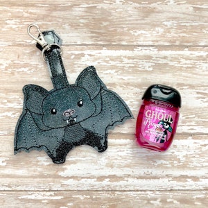 May include: A black bat-shaped hand sanitizer holder with a silver keychain. The holder is made of fabric and has a sparkly finish. The holder is next to a pink hand sanitizer bottle with the text "Ghoul Friend" and a pumpkin graphic.