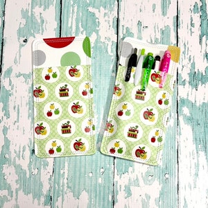 May include: Two rectangular pen holders with a white, red, and green apple pattern. One holder contains pens and markers. The top section features a polka dot design. The edges are stitched with white thread. The background is a distressed, light blue wooden surface.